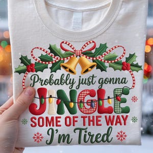 Op de afbeelding: Crèmekleurig T-shirt met een feestelijk kerstontwerp. Het ontwerp toont de woorden "Probably just gonna JINGLE SOME OF THE WAY I'm Tired" in rood, groen en crème, met hulst, bellen en lichtslingers.