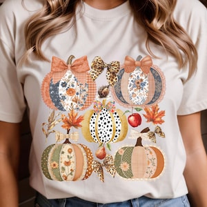 Op de afbeelding: Een crèmekleurig T-shirt met een herfstthema. Het ontwerp bevat zes decoratieve pompoenen met verschillende patronen, strikken en herfstbladeren. De pompoenen zijn in tinten oranje, groen en goud.