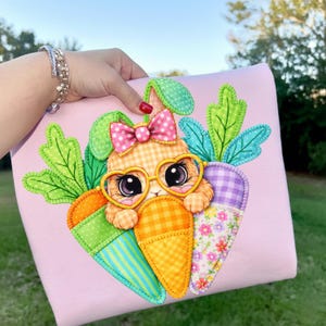 May include: A pink pillow featuring a cartoon bunny wearing heart-shaped glasses and a pink bow. The bunny is nestled within a carrot design with green, orange, and purple accents. The pillow is held by a person.