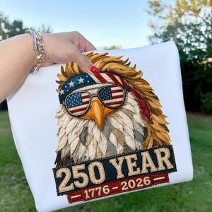 May include: White sweatshirt with an eagle design wearing sunglasses with an American flag pattern. The eagle has a red, white, and blue headband. The text "250 YEAR 1776-2026" is displayed below the eagle in a banner.