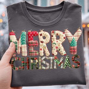 May include: A dark grey crew neck t-shirt featuring the words "MERRY CHRISTMAS" appliquéd across the front. The letters are crafted from diverse patterned fabrics, including plaid and polka dots, and are outlined with gold beads. The shirt is held up by a hand.