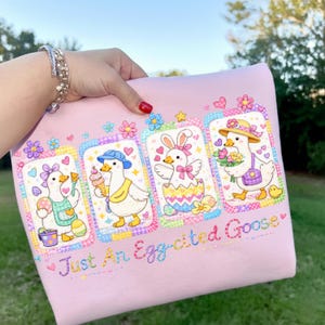 May include: Pink fabric featuring four cartoon geese in decorative frames. Each goose is dressed up with accessories like a hat, bow, or apron. The text "Just An Egg-cited Goose" is embroidered in a cursive font. The design includes hearts, stars, and flowers.