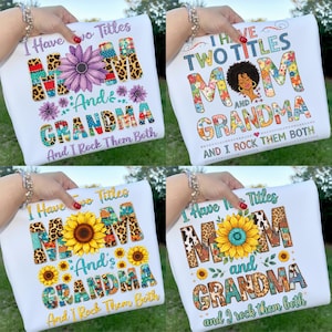 Op de afbeelding: Witte T-shirts met de tekst "I Have Two Titles MOM and GRANDMA And I Rock Them Both". Het woord "MOM" is versierd met bloemen-, luipaard- en patchworkpatronen. Het woord "GRANDMA" is versierd met bloemen- en patchworkpatronen. De shirts hebben een zonnebloem of een paarse bloem.