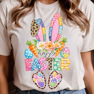 May include: A cream-colored t-shirt featuring a colorful Easter design. The design includes bunny ears, carrots, Easter eggs, flowers, and the words "Happy Easter". The bunny ears and Easter eggs have patchwork patterns.
