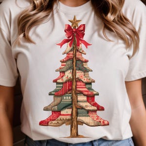 May include: Cream-colored t-shirt featuring a Christmas tree design made of stacked cowboy boots. The boots are red, green, and beige with various patterns. A red bow and gold star adorn the top of the tree.