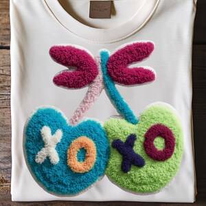 May include: White t-shirt with a colorful, textured cherry design. The cherries are teal and green, with the letters "XOXO" in white, orange, and purple. The stem and dragonfly wings are pink, blue, and magenta.