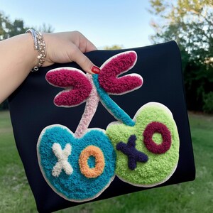 May include: Black clutch with a colorful, textured design featuring a dragonfly and two heart-shaped cherries. The cherries are teal and green, with white, orange, and purple accents. The dragonfly is pink and blue.