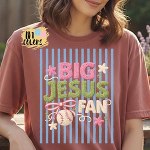 Puede incluir: Camiseta de color rosa polvoriento con la frase "BIG JESUS FAN" en rosa, verde y blanco. El diseño incluye una pelota de béisbol, flores y rayas verticales azules. La camiseta también tiene el texto "In Colors" en la esquina superior izquierda.