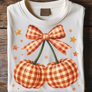 May include: Cream-colored t-shirt featuring a whimsical design of two pumpkins connected by green stems and a plaid bow. The pumpkins and bow have orange and white checkered patterns. Scattered orange stars complete the fall-themed graphic.
