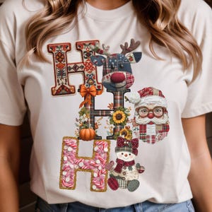 Op de afbeelding: Crèmekleurig T-shirt met een feestelijk ontwerp. De letters "H O H O" worden weergegeven in patchwork-stijl. De "H"'s zijn versierd met bloemen en patronen, en de "O"'s tonen een rendier, de kerstman en een sneeuwpop.