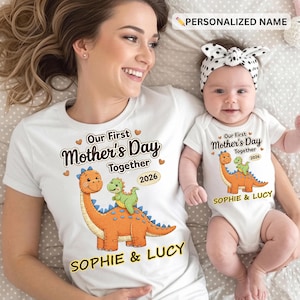 May include: White t-shirts and a baby bodysuit with a dinosaur graphic. The shirts read "Our First Mother's Day Together 2026" and "SOPHIE & LUCY". The baby wears a white bodysuit with a matching design. The adult is smiling.
