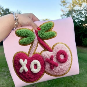 Könnte beinhalten: Ein hellrosa Kissen mit einem getufteten Kirschdesign. Die Kirschen sind in Rosa- und Rottönen gehalten, mit grünen Blättern und goldenen Verzierungen. Das Design enthält die Buchstaben "XO" in Weiß und Rot.