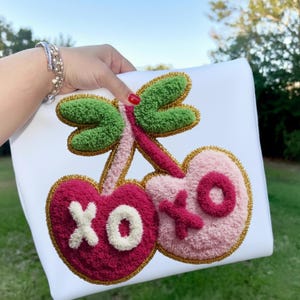 May include: White pillow with a textured design of two cherries and a dragonfly. The cherries are pink and red with the letters "XO" in white. The dragonfly has green wings and a pink body. Gold trim outlines the design.