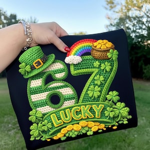 Könnte beinhalten: Schwarzes T-Shirt mit einem Häkeldesign mit der Zahl 67, einem Koboldhut, einem Regenbogen, einem Goldtopf und Kleeblättern. Das Wort "LUCKY" wird auf einem Banner angezeigt. Goldmünzen sind unten verstreut.