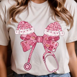 以下が含まれることがあります： ピンク色のクリスマスをテーマにしたデザインが施された明るい色のTシャツ。デザインには、サンタの帽子2つ、リボン、聴診器が含まれています。リボンと帽子は、クリスマスのアイコンが入ったチェック柄です。聴診器はピンク色です。