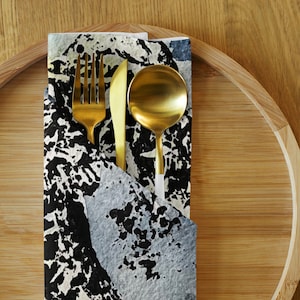 Könnte beinhalten: Ein goldfarbenes Gabel-, Messer- und Löffel-Set auf einer Serviette mit einem abstrakten Design in Schwarz, Weiß und Blau. Die Serviette liegt auf einem Holztablett und schafft eine stilvolle Tischdekoration.