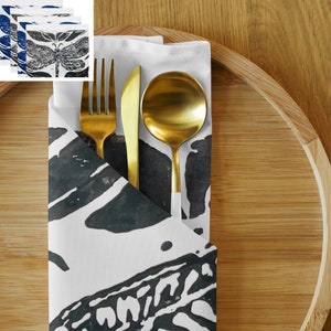 May include: A folded white napkin with a black and white dragonfly print, holding a gold fork, knife, and spoon. The utensils are arranged on a bamboo tray, creating a stylish table setting. Additional napkins with dragonfly prints are visible.