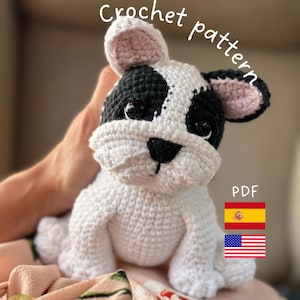 Puede incluir: Un perro de ganchillo hecho a mano en blanco y negro, con el texto "Crochet pattern" y "PDF". El perro tiene orejas internas rosas y está sostenido por una persona. La imagen también incluye las banderas de España y Estados Unidos.