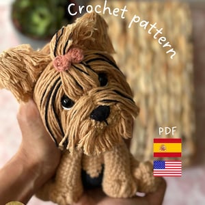 Puede incluir: Un perro Yorkshire Terrier tejido a ganchillo con hilo marrón y detalles negros. El perro tiene un lazo rosa y está etiquetado como "Crochet pattern" y "PDF". La imagen incluye las banderas española y estadounidense.