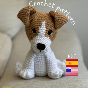 Puede incluir: Un juguete de perro tejido a ganchillo en blanco y marrón, con el texto "Crochet pattern" y "PDF". El perro tiene ojos negros y una nariz negra. La imagen también incluye las banderas española y estadounidense.