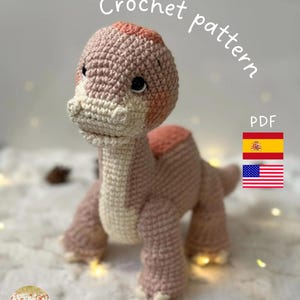 Puede incluir: Un juguete de dinosaurio tejido a ganchillo en tonos rosa y crema, con las palabras "Crochet pattern" y "PDF" en blanco. La imagen también incluye las banderas de España y Estados Unidos.
