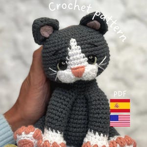 Puede incluir: Un gato de ganchillo hecho a mano en gris, blanco y melocotón. El gato tiene pecho, patas y hocico blancos, con detalles en color melocotón en las patas y la nariz. Las palabras "Crochet pattern" y "PDF" son visibles. La imagen también incluye las banderas de España y Estados Unidos.