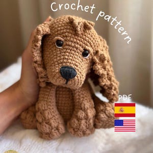 Puede incluir: Patrón de crochet para un perro amigurumi marrón. El patrón está disponible en formato PDF e incluye instrucciones en inglés y español. El perro está sentado con la cabeza inclinada hacia un lado y tiene una expresión linda.