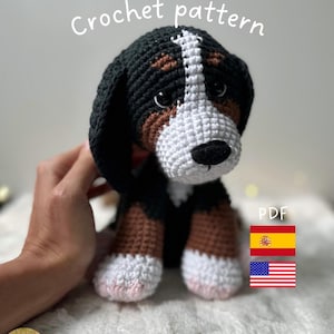 Puede incluir: Un cachorro de perro de montaña bernés tejido a ganchillo con hilo negro, marrón y blanco. El cachorro tiene una raya blanca en la cara y detalles rosas en las patas. Las palabras "Crochet pattern" están escritas encima del cachorro. La imagen también incluye las letras "PDF" y las banderas de España y Estados Unidos.