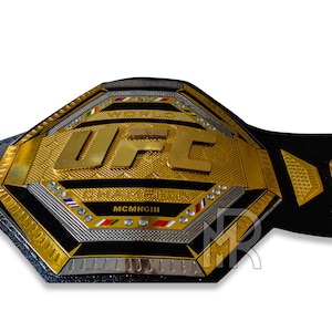 May include: A gold and black UFC World Championship belt with the UFC logo and the year MCMXCIII. The belt is made of leather and has a metal buckle.
