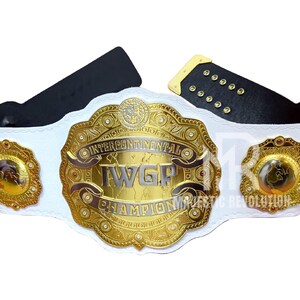 May include: A white and gold wrestling championship belt with the text "Intercontinental IWGP Champion". The belt has a large central plate and two smaller side plates, all with intricate gold detailing. The belt's strap is white with black leather backing.