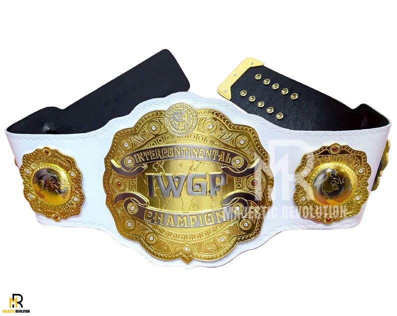 May include: A white and gold wrestling championship belt with the text "Intercontinental IWGP Champion". The belt has a large central plate and two smaller side plates, all with intricate gold detailing. The belt's strap is white with black leather backing.