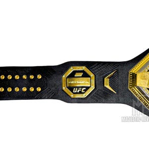 May include: A black leather UFC championship belt with gold accents and the UFC logo. The belt has a hexagonal buckle with the UFC logo and the words "UFC" and "World Championship" inscribed on it.