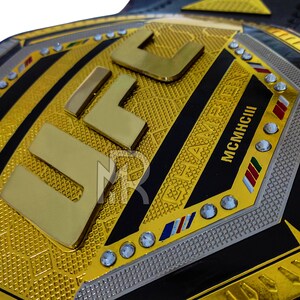 May include: A gold UFC championship belt with black and silver accents. The belt features the UFC logo and the words "CHAMPION" and "MCMXCIII" in gold lettering. The belt is adorned with rhinestones.