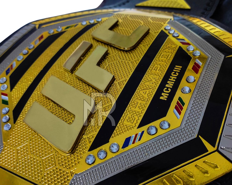 May include: A gold UFC championship belt with black and silver accents. The belt features the UFC logo and the words "CHAMPION" and "MCMXCIII" in gold lettering. The belt is adorned with rhinestones.
