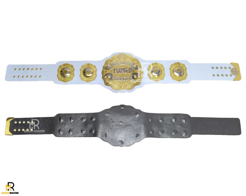 May include: Two wrestling championship belts. The top belt is white with gold accents and a central gold emblem that reads "IWGP CHAMPION". The bottom belt is dark gray with a textured surface and gold accents.