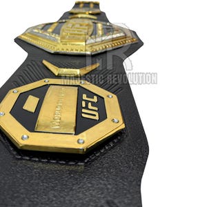 May include: A black leather UFC championship belt with gold accents. The belt features a large octagon-shaped plate with the words "UFC" and "WORLD CHAMPION" inscribed on it.