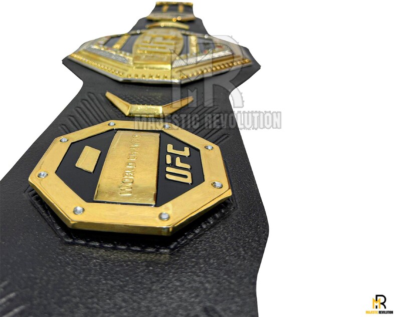 May include: A black leather UFC championship belt with gold accents. The belt features a large octagon-shaped plate with the words "UFC" and "WORLD CHAMPION" inscribed on it.