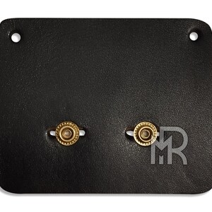 May include: A black leather patch with two gold-colored metal rings. The rings have a decorative pattern around the edge.