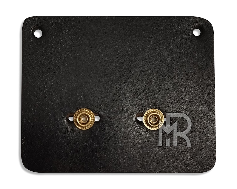 May include: A black leather patch with two gold-colored metal rings. The rings have a decorative pattern around the edge.