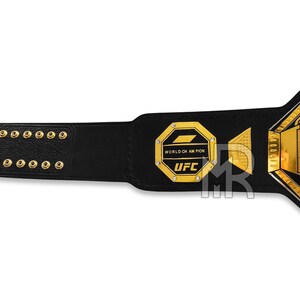 May include: A black leather UFC championship belt with gold accents. The belt features a large octagon-shaped plate with the words "WORLD CHAMPION" and "UFC" in gold lettering.