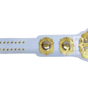 May include: A white wrestling championship belt with gold accents. The belt features a large central plate with the words "Intercontinental IW" and two smaller side plates. The strap has multiple holes for adjustment.