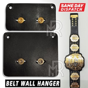 May include: Two black leather belt wall hangers with gold metal hooks. The hangers are designed to hold belts vertically.  BELT WALL HANGER is printed in white letters on the bottom of the image.