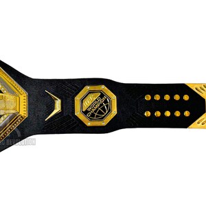 May include: A gold and black UFC World Champion belt with a black leather strap and gold accents. The belt features a large octagon-shaped buckle with the UFC logo and the words "UFC World Champion".