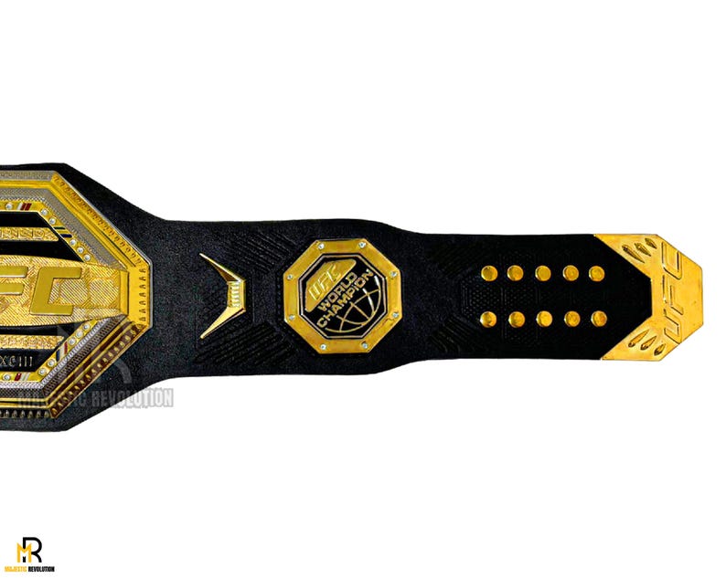 May include: A gold and black UFC World Champion belt with a black leather strap and gold accents. The belt features a large octagon-shaped buckle with the UFC logo and the words "UFC World Champion".