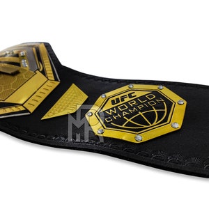 May include: A black and gold UFC World Champion belt with a hexagonal plate featuring the UFC logo and the words "UFC World Champion".