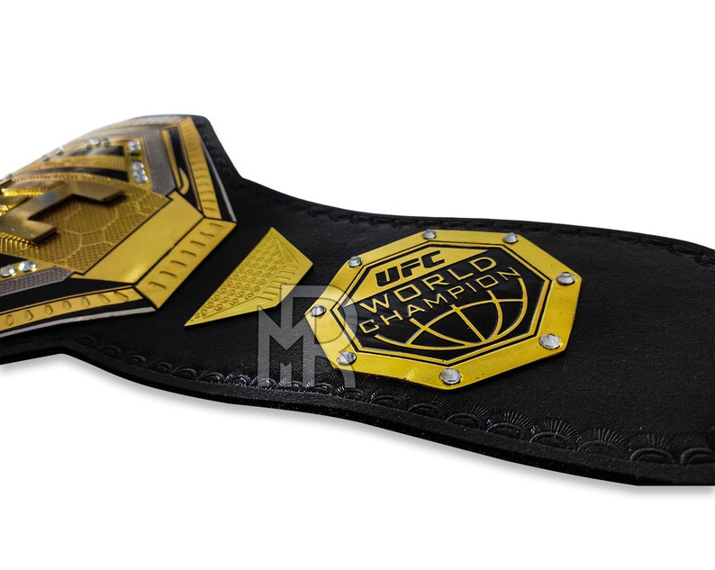 May include: A black and gold UFC World Champion belt with a hexagonal plate featuring the UFC logo and the words "UFC World Champion".