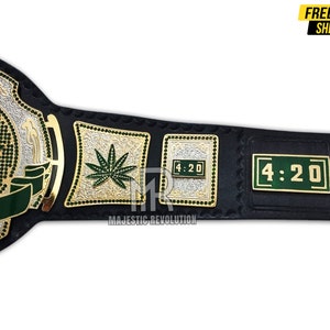 Pode incluir: Um cintur&atilde;o de campeonato de luta livre de couro preto com detalhes dourados e prateados. O cintur&atilde;o apresenta um design de folha de maconha verde, as palavras "4:20" e o texto "Majestic Revolution".
