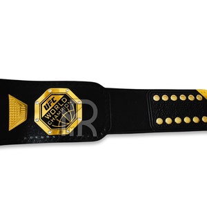 May include: A black leather UFC World Champion belt with a gold buckle and gold accents. The buckle features the UFC logo and the words "World Champion".