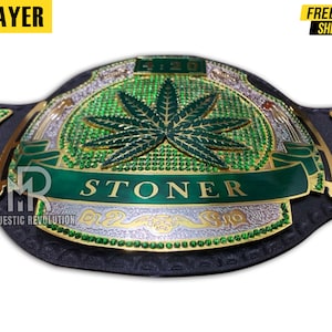 Pode incluir: Um cintur&atilde;o de campeonato verde e dourado com um design de folha de maconha e a palavra "STONER" em uma faixa verde. O cinto &eacute; feito de couro e tem uma fivela dourada.