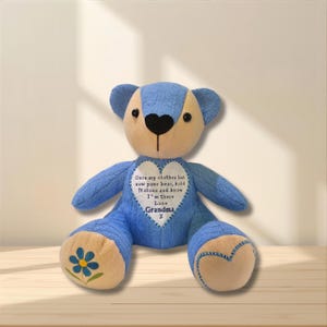 May include: A blue and beige teddy bear featuring a heart-shaped patch on its chest with the text: Once my clothes but now your bear, hold it close and know I'm there Love Grandma x. The bear has a blue flower embroidered on its left foot.